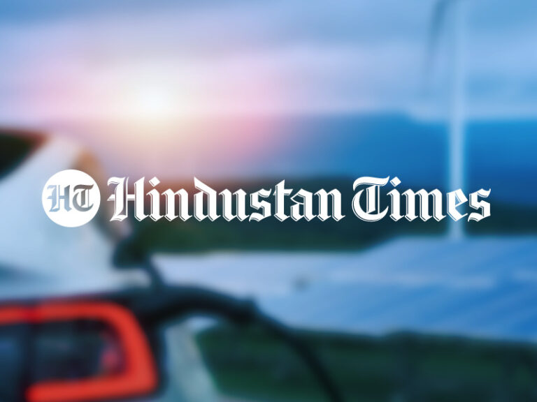 The Hindustan Times logo over a blurred photo of an electric vehicle charging and solar panels and wind turbines in the background